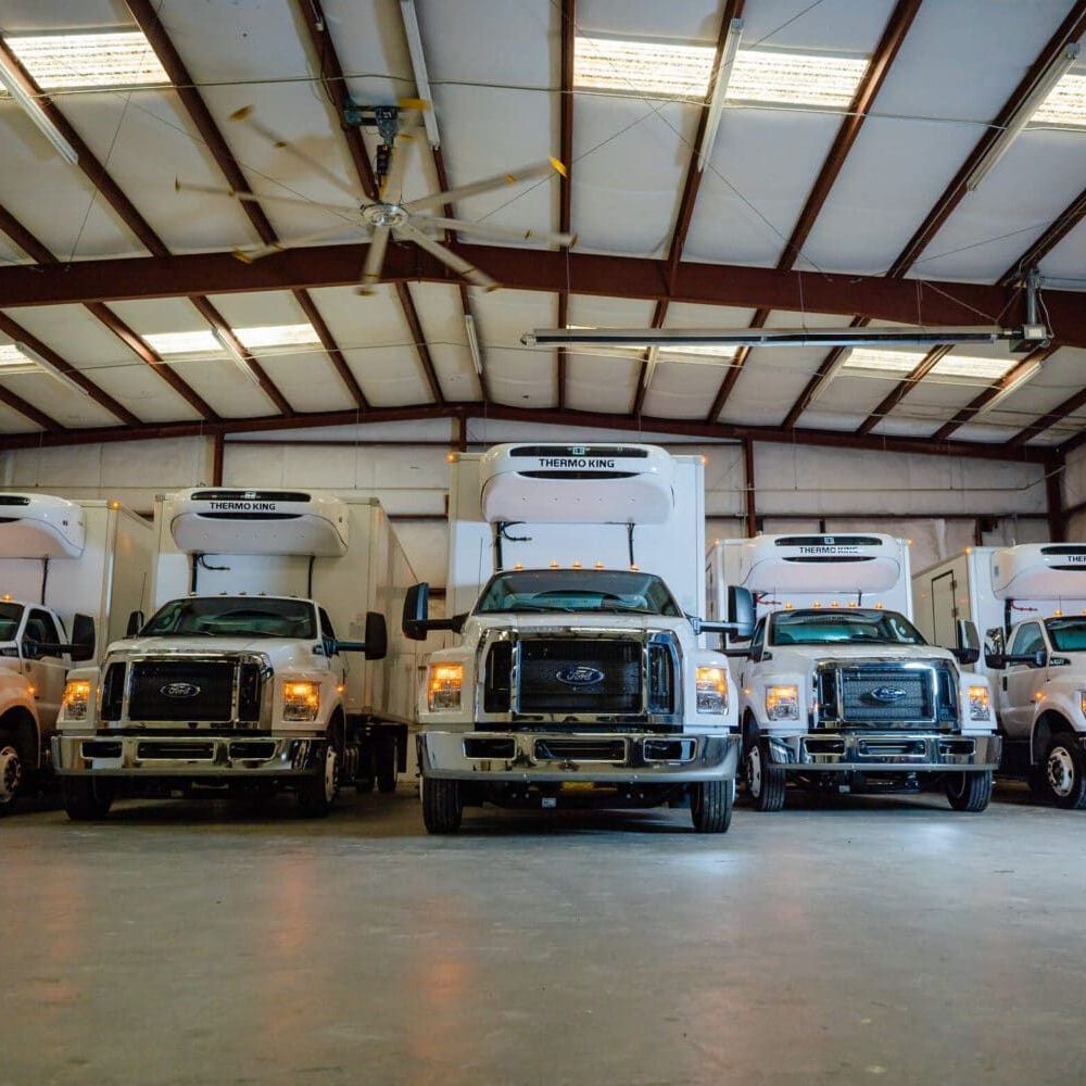 A row of trucks are parked in a warehouse under a ceiling fan