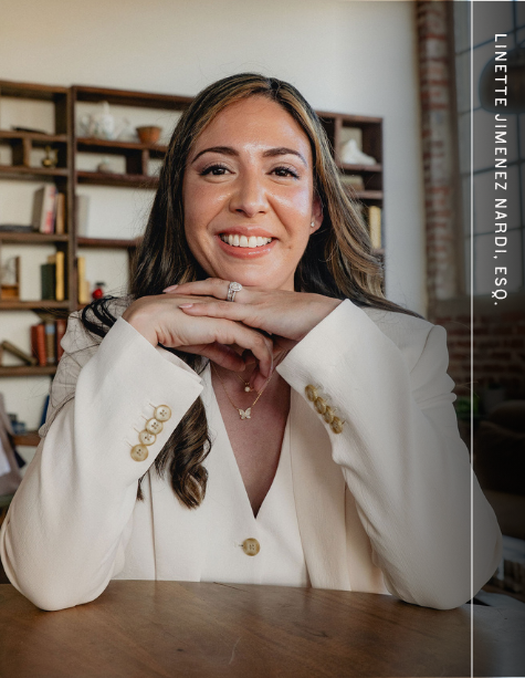 Smiling woman in white blazer seated at a table, hands under chin, with shelves in the background