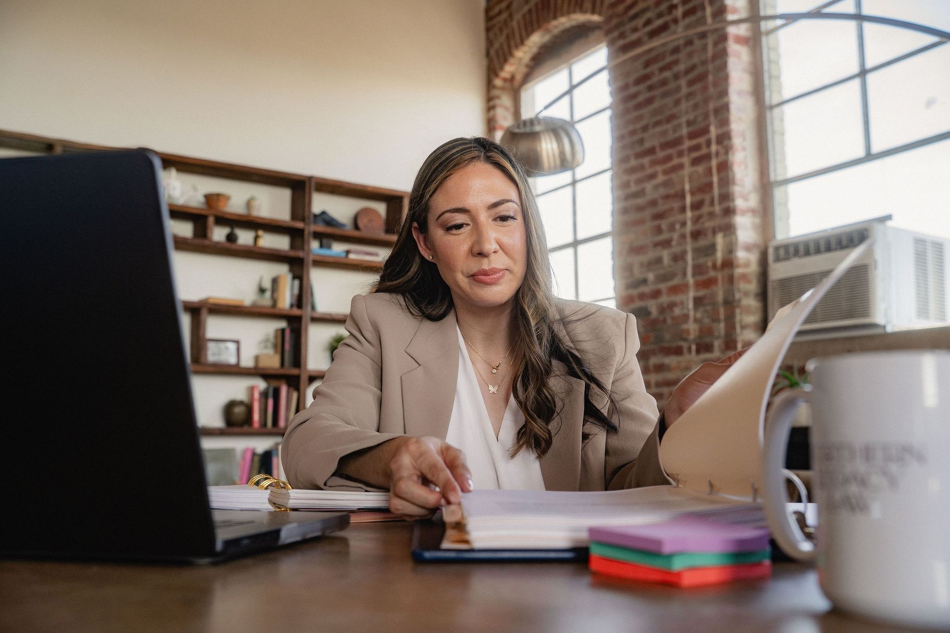 Woman working at a desk with laptop, papers, and notebook in a bright office