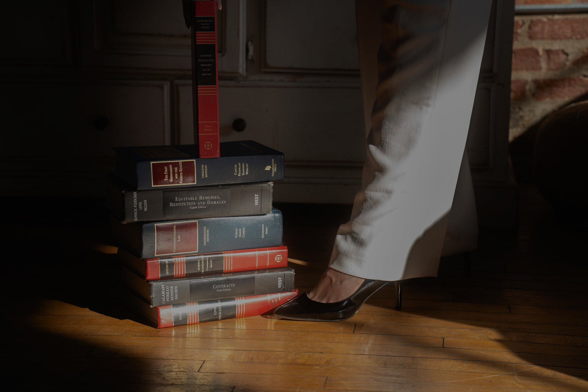 A person in white pants steps near a stack of books on a wooden floor in dim light