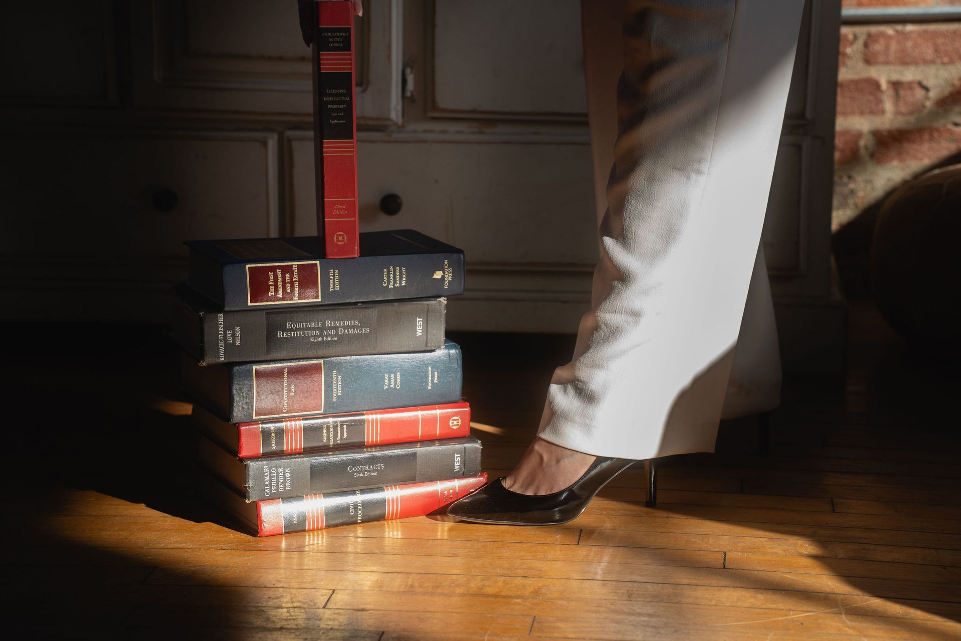 Stack of books beside a person’s white-clad leg and black shoe on a wooden floor, lit by warm sunlight
