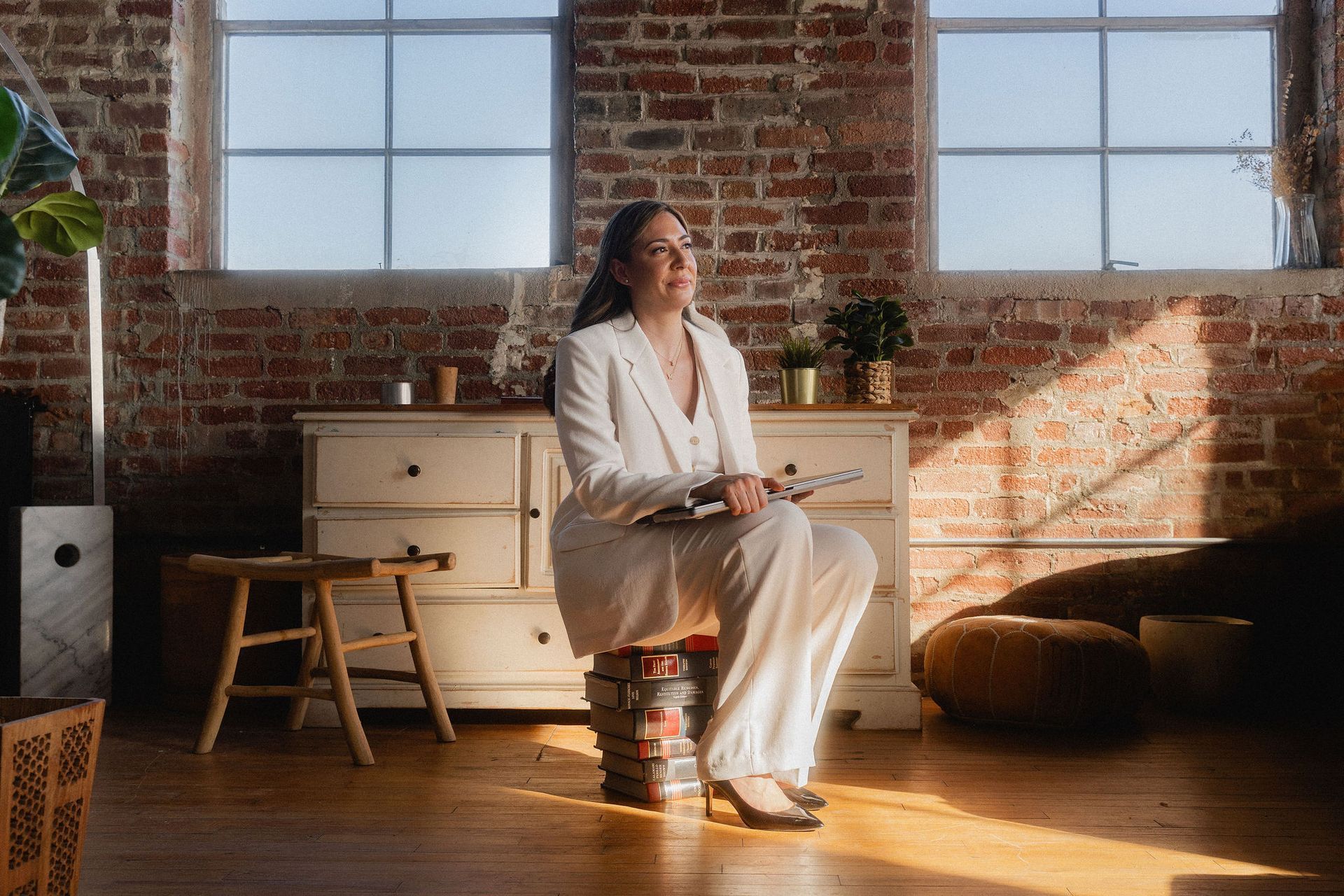 Person seated by a sunlit brick wall in a cozy room, wearing a white suit and heels.