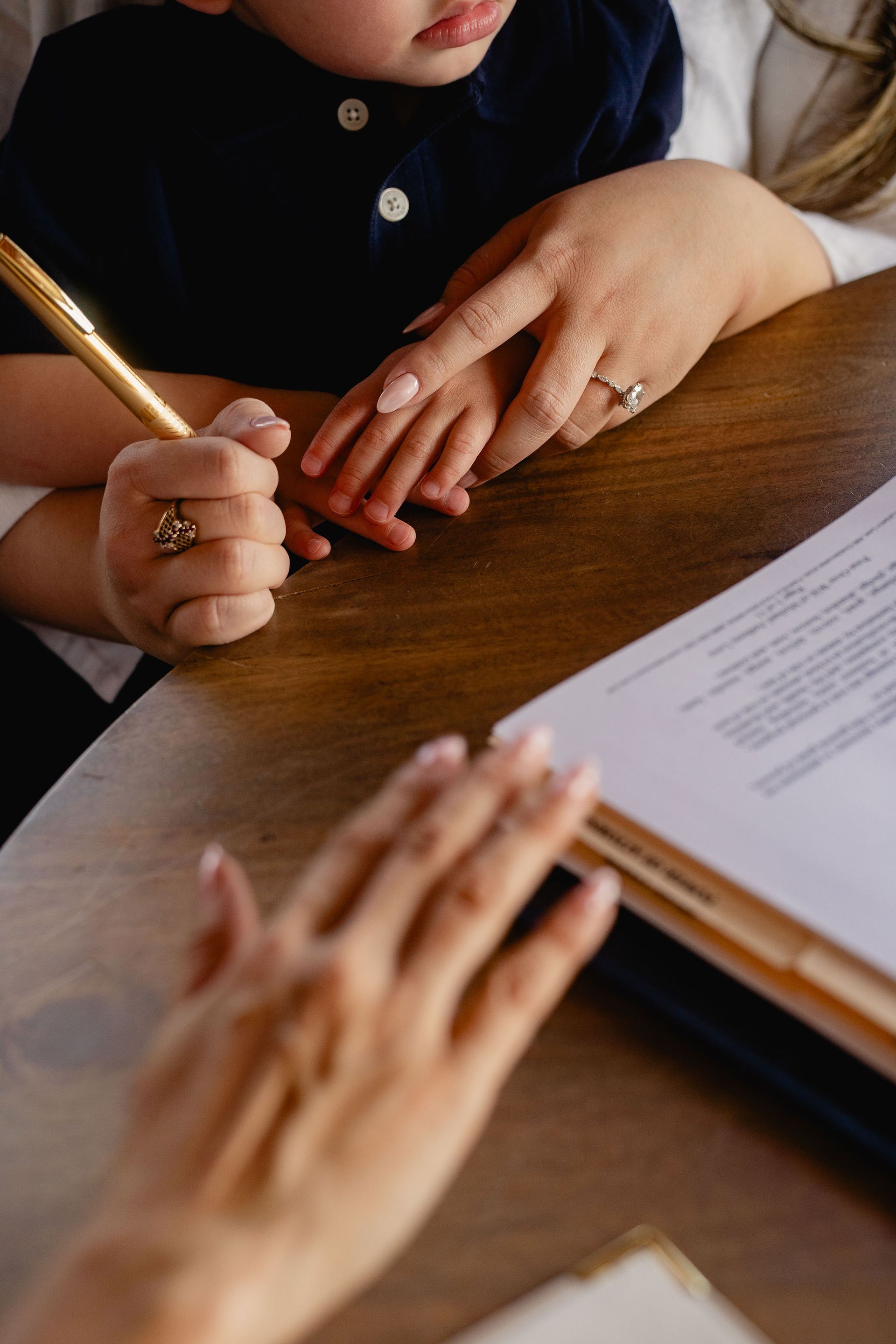 Hands guiding a child’s pencil as they write at a wooden table beside a notebook.