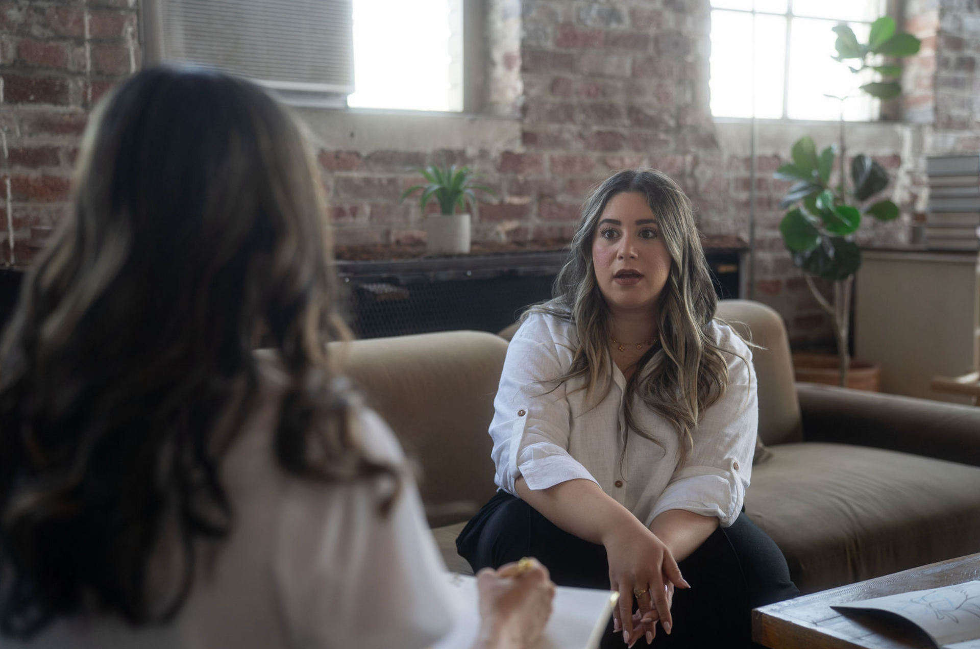 Two people talking on a couch in a bright room with exposed brick walls and plants.