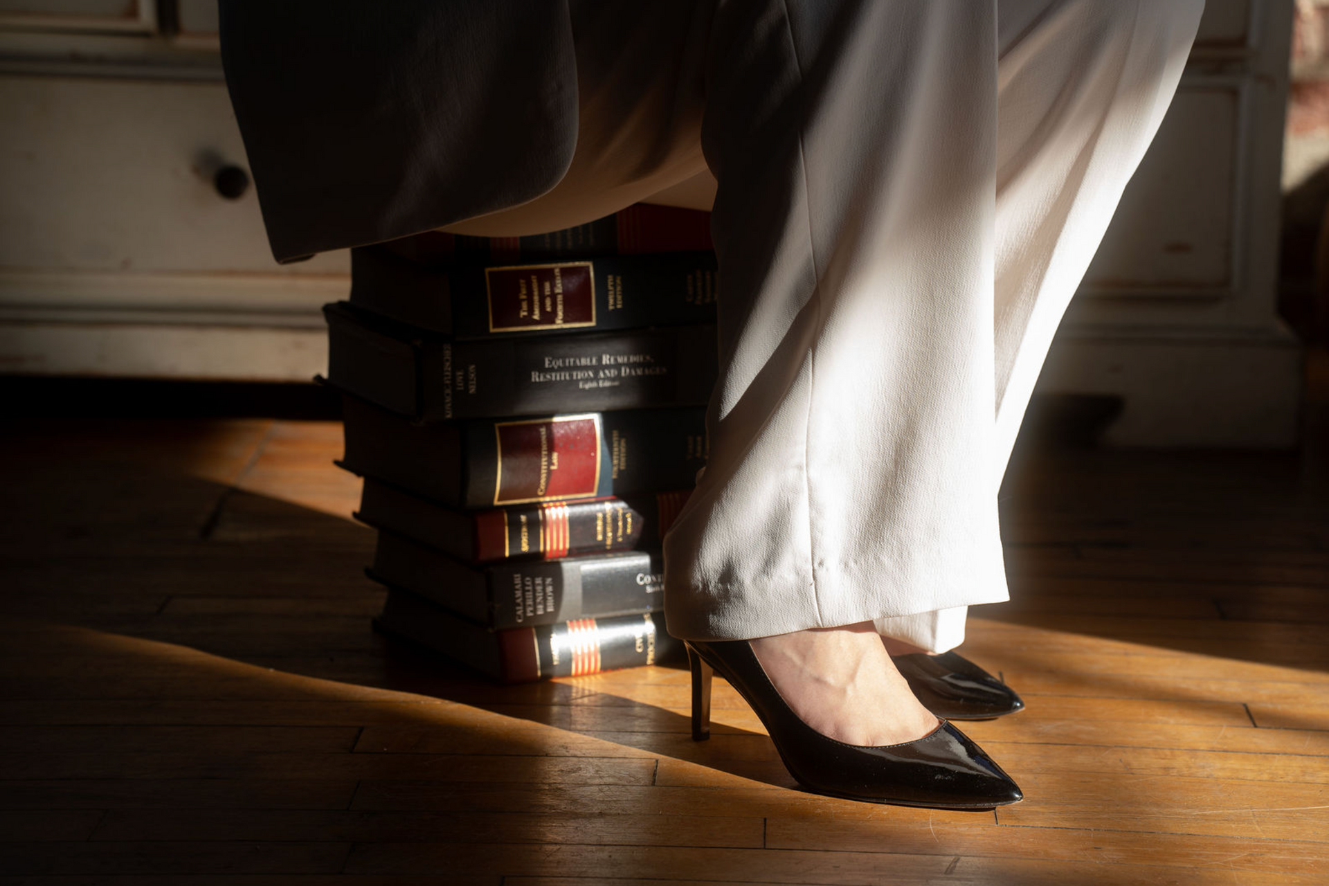 Woman’s feet on a stack of books, wearing black heels and white pants on a wooden floor.