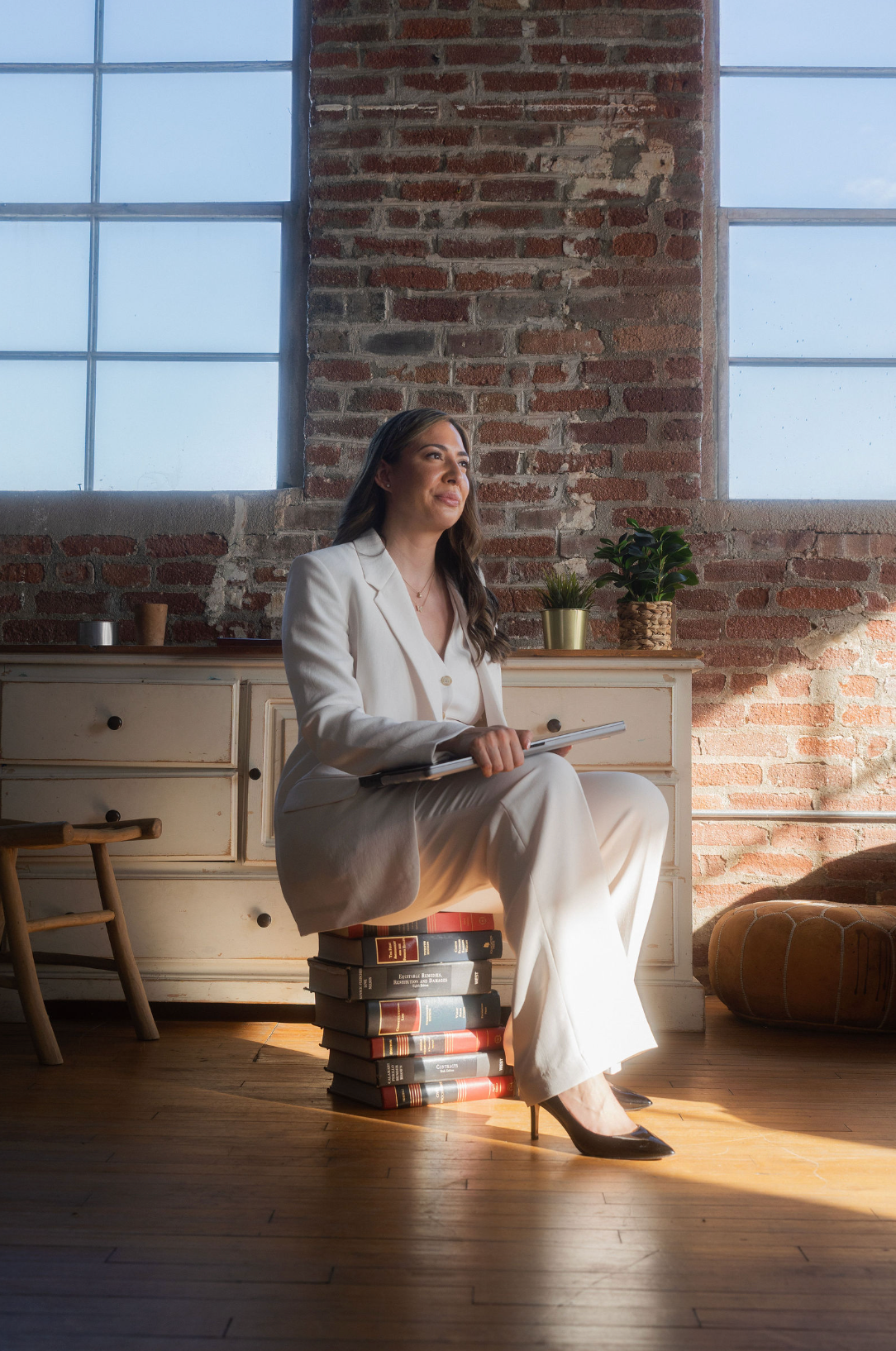 Woman seated on a stack of books in a sunlit loft, holding a laptop near large windows.