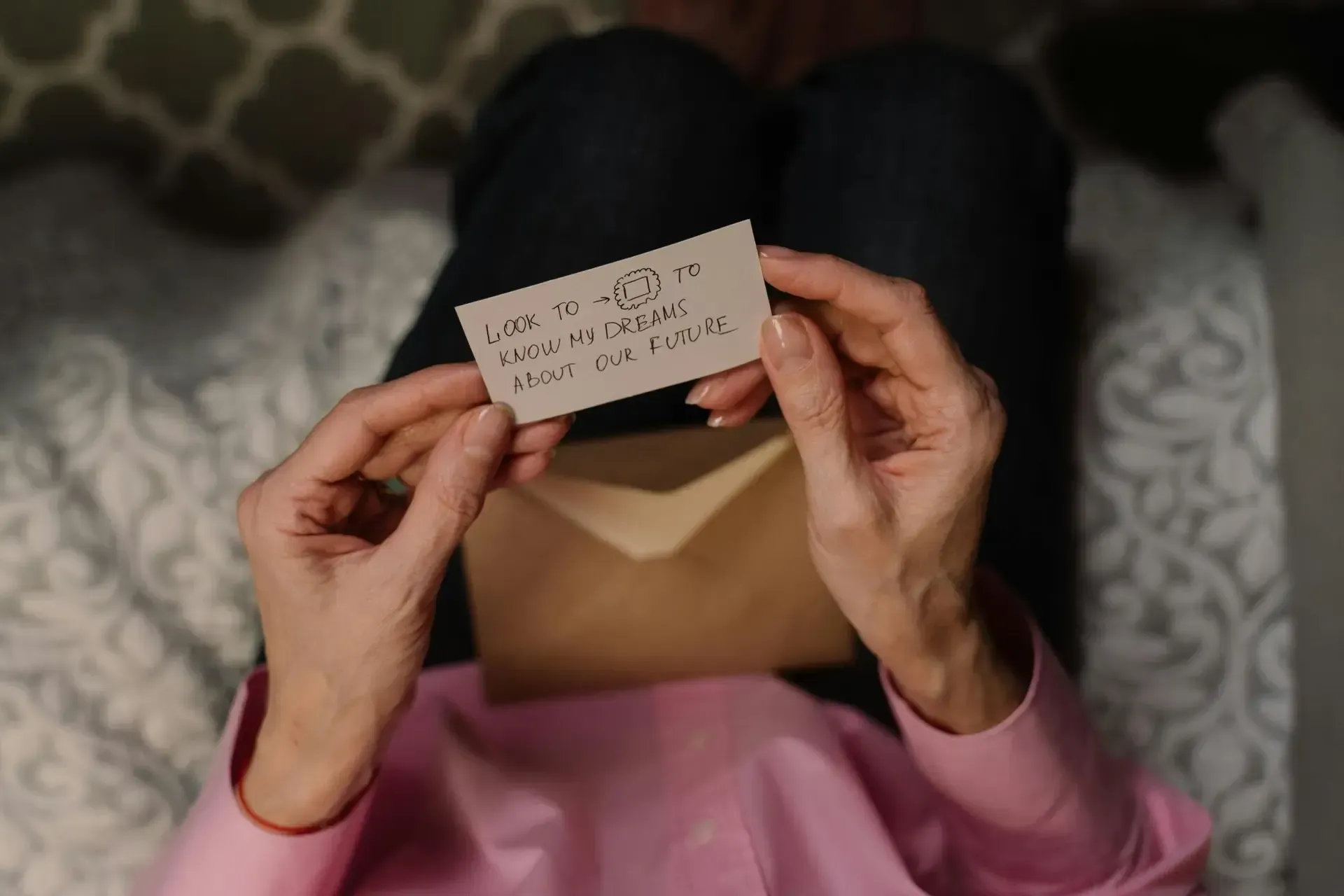 Person in pink shirt holding a handwritten note over an open envelope on a patterned bedspread