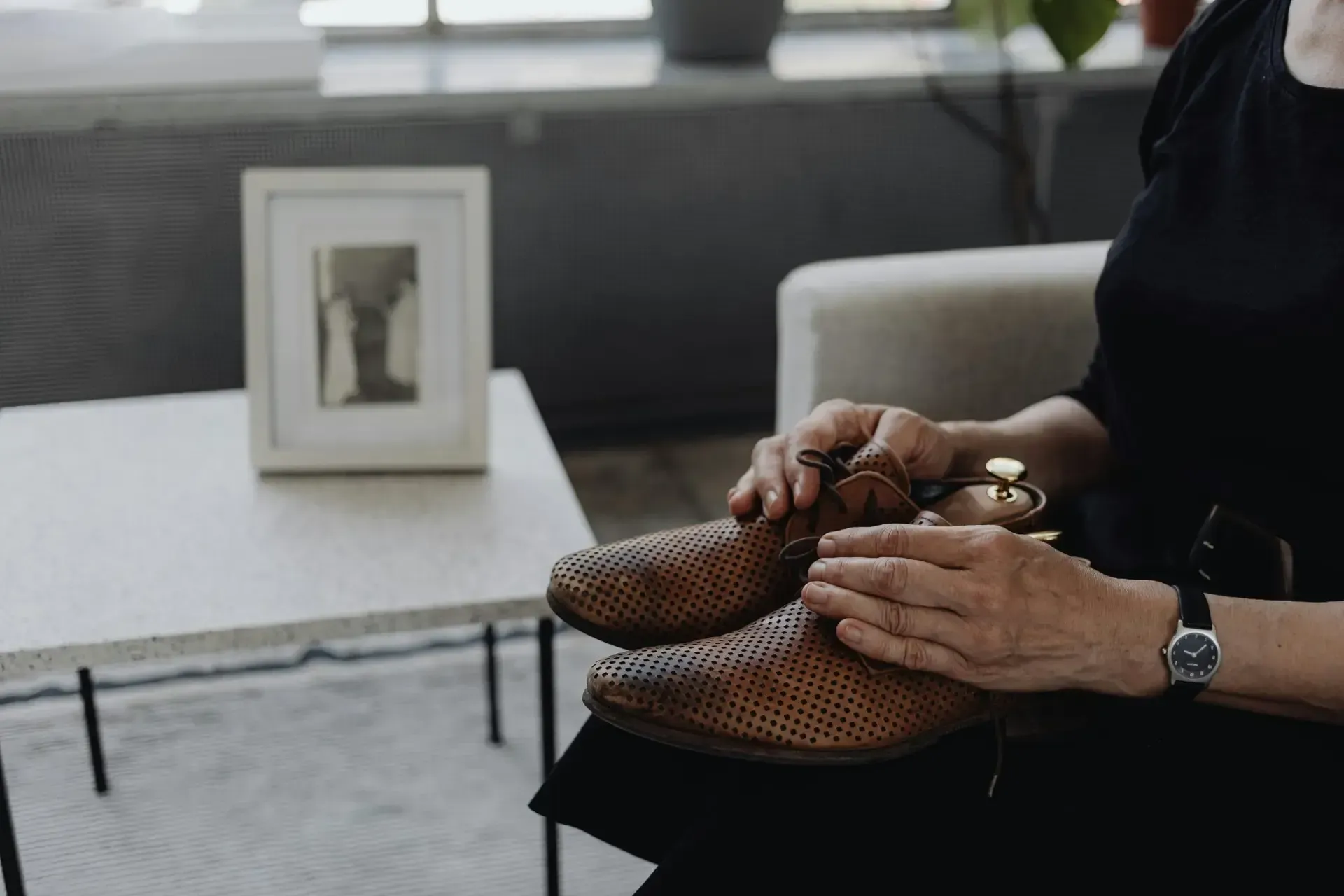 Hands hold a pair of brown woven leather shoes while sitting in a room with a framed photo on a side table.