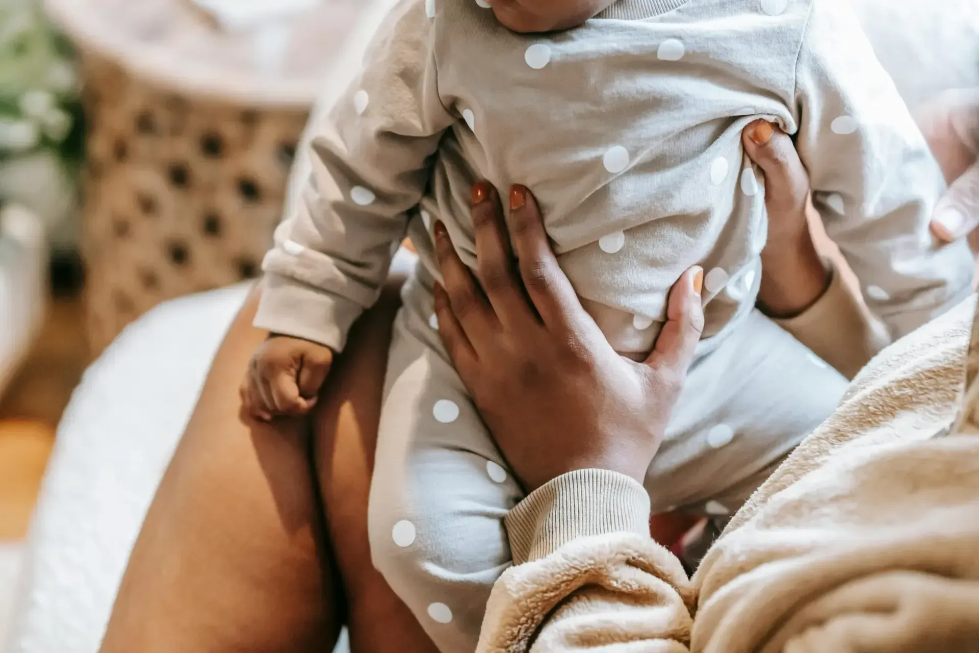 A person in a beige sweater holds a baby wearing a beige and white polka-dot outfit on their lap.