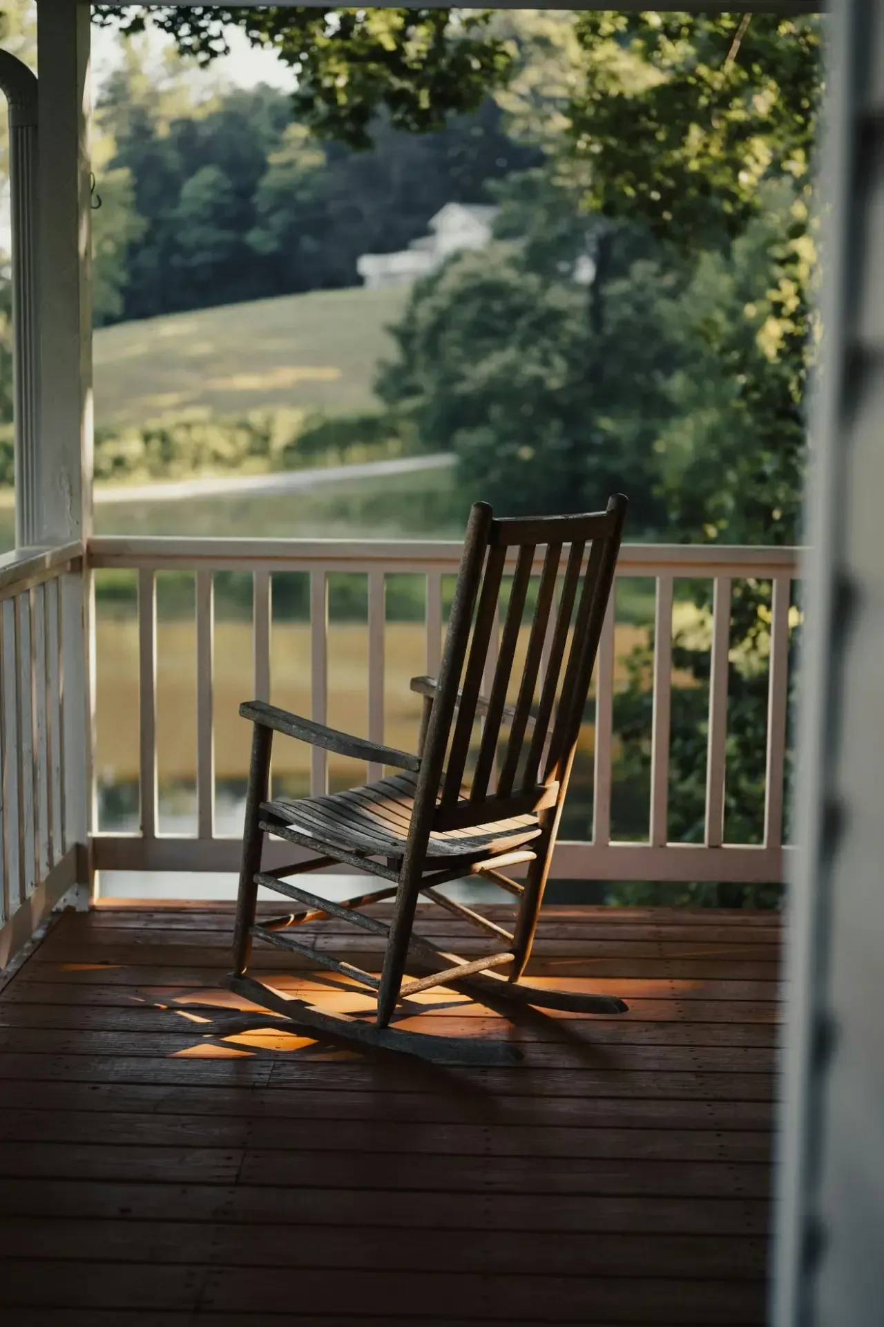 A wooden rocking chair sits on a porch overlooking a serene landscape with rolling green hills and a small pond at sunset.