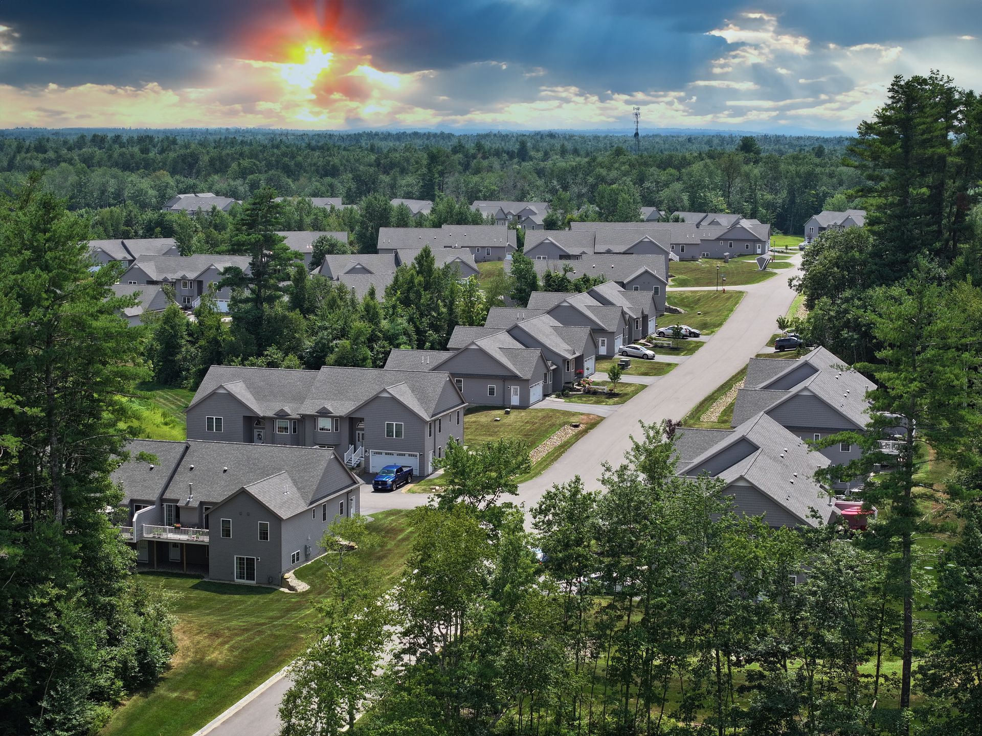 Aerial view of a 55+ neighborhood with gray houses, green trees, and a bright sunset sky.