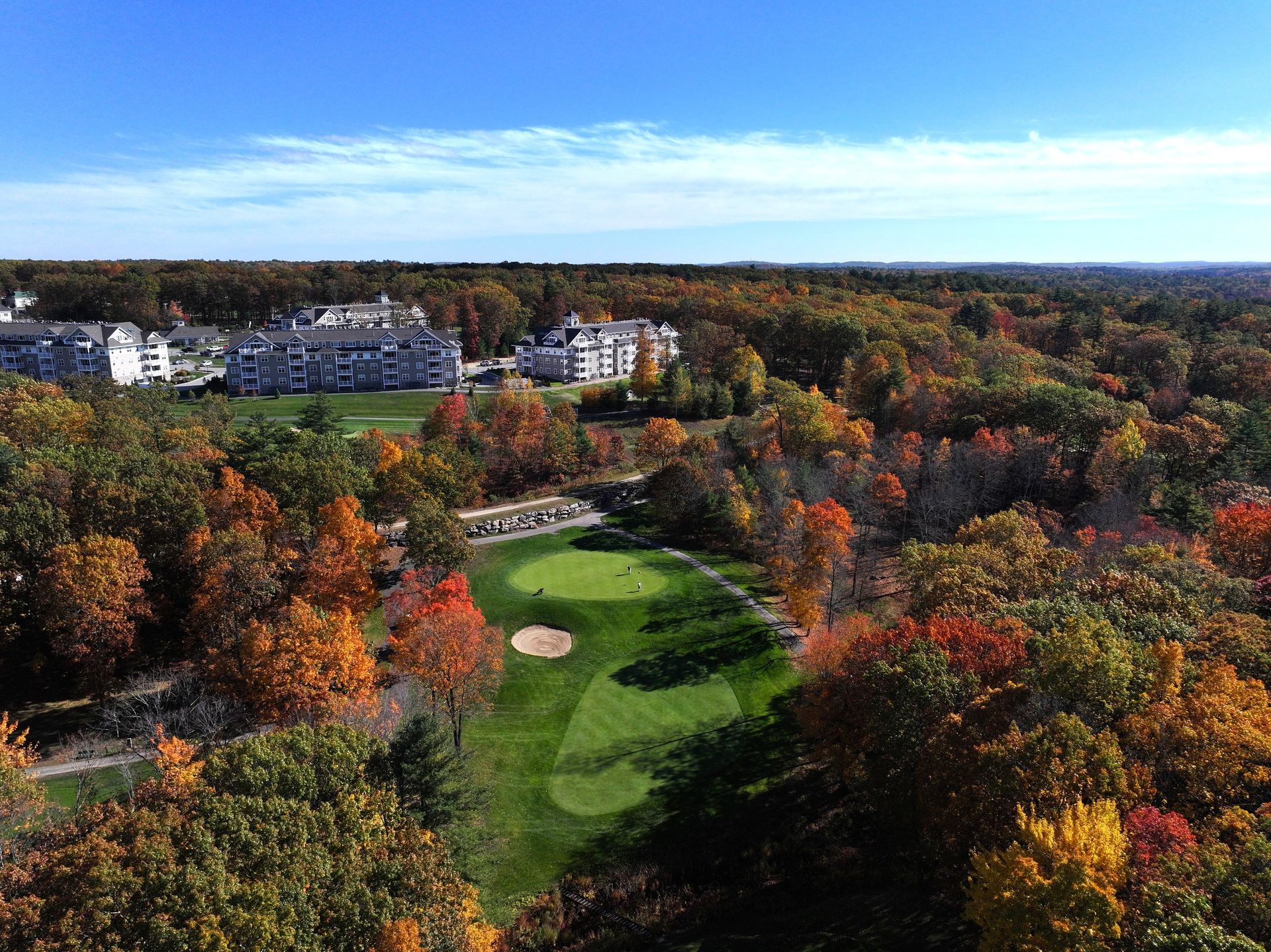 Autumn golf course surrounded by colorful trees and nearby buildings under a clear sky