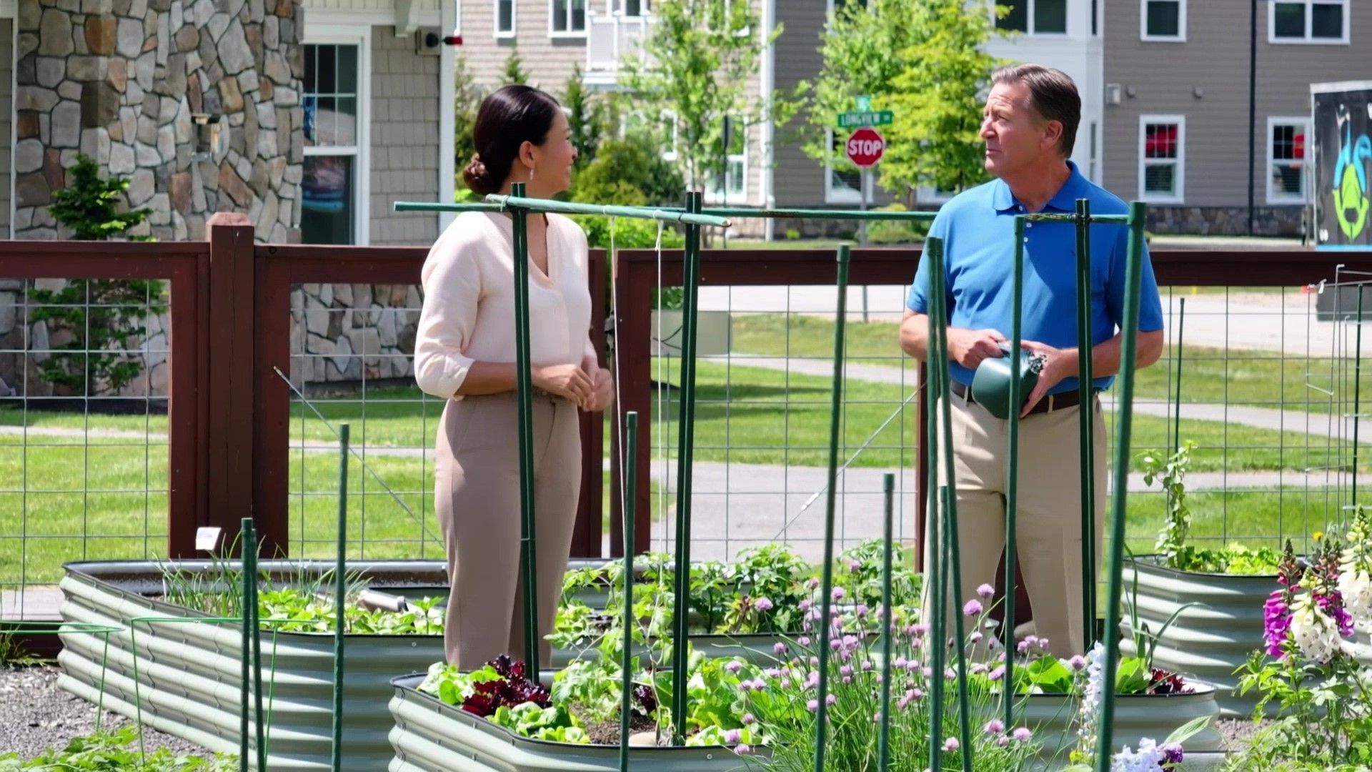 Two people talk in a community garden with raised beds and luxury condominiums behind them.