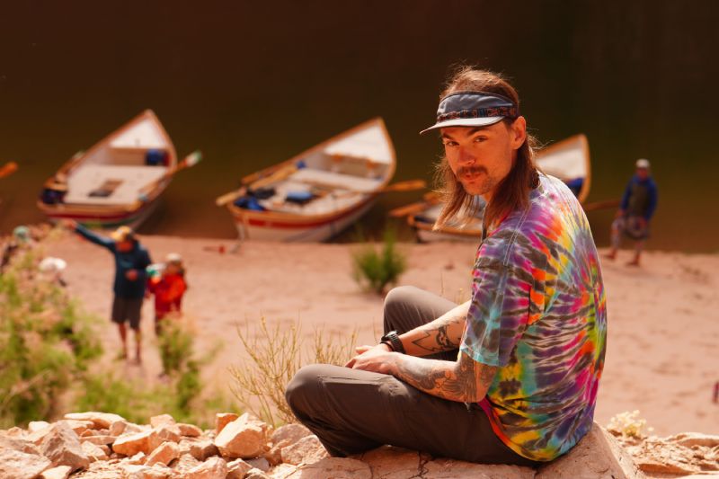 A man in a tie dye shirt is sitting on a rock with boats in the background.