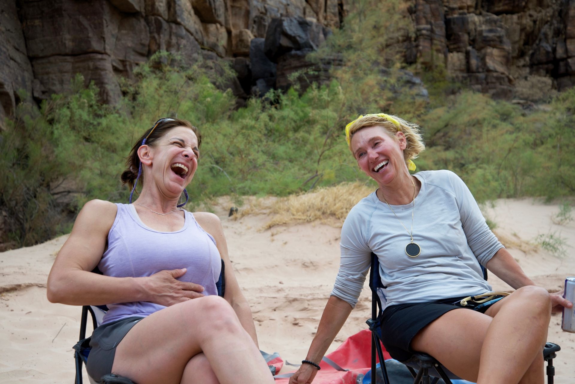 Two women sit in chairs on the beach laughing