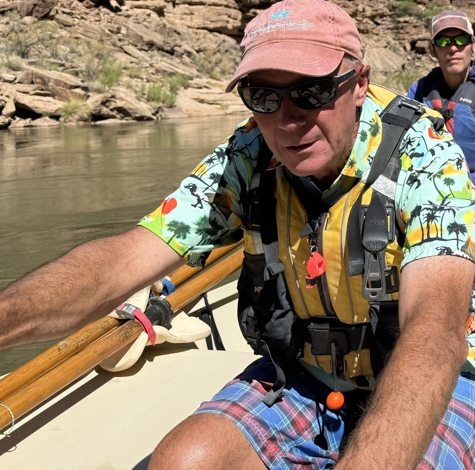 Man in cap and sunglasses rows a raft, wearing a life vest and colorful shirt. River setting.