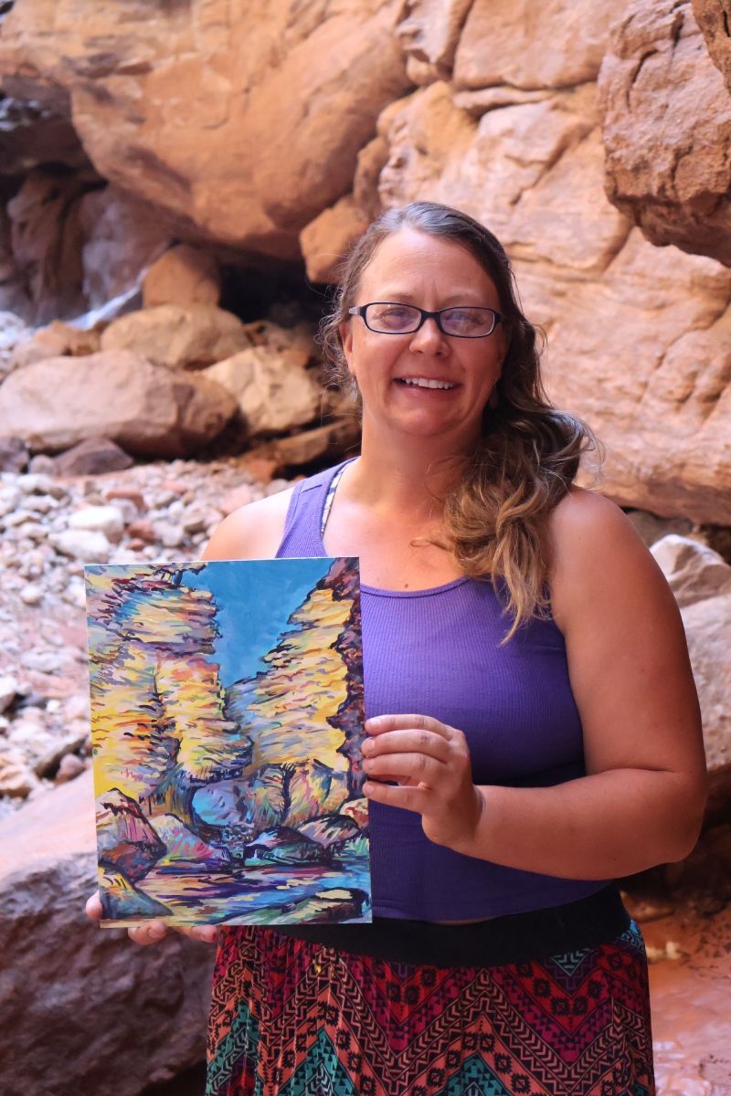 A woman is holding a painting in front of a rock wall.