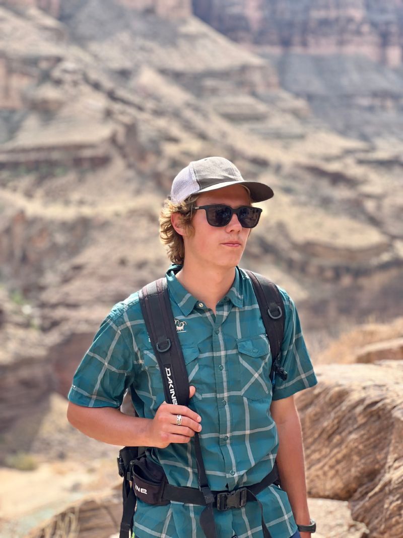 A young man wearing sunglasses and a hat is standing in front of a mountain.