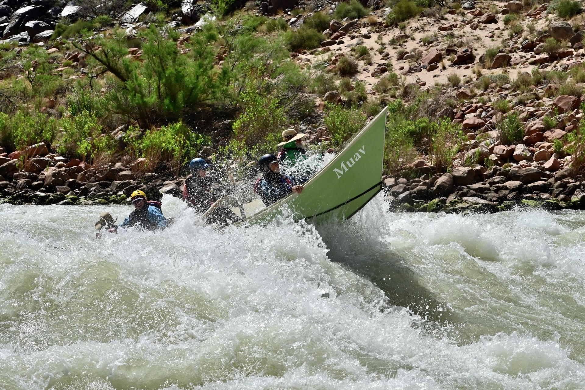 Kayaker in a green boat capsizing in rough whitewater beside a rocky shore