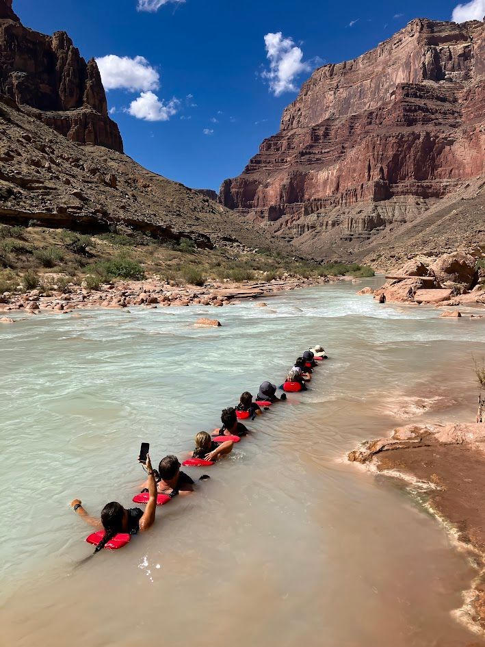 Group of people floating in a river with red canyon walls and blue sky.