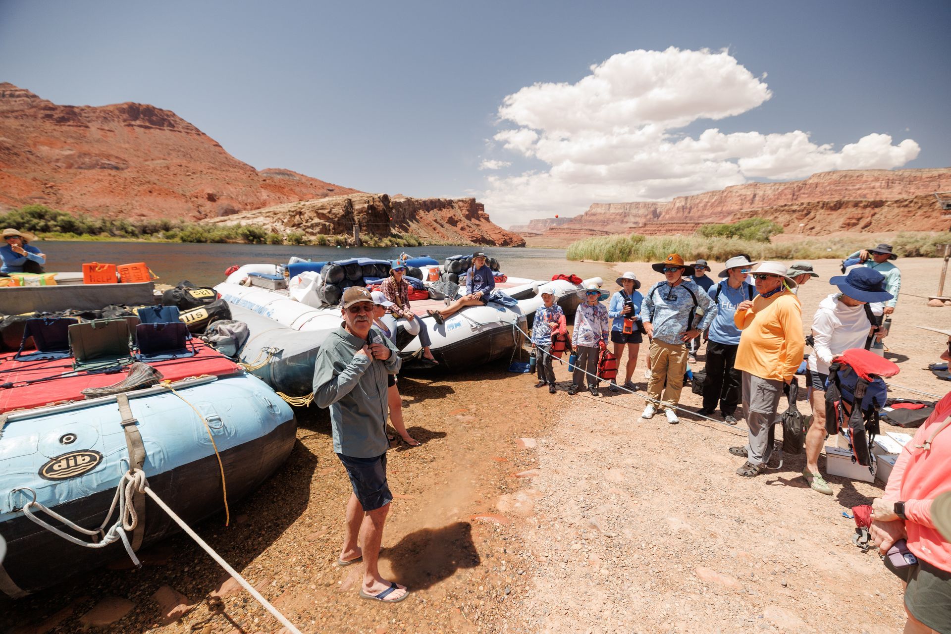 People listen to a guide near rafts on a riverbank, red rock canyon in background under a sunny sky.