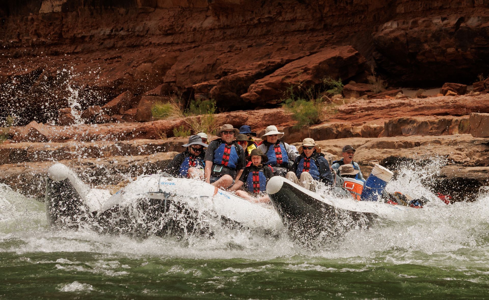 Whitewater rafters in colorful helmets splash through rapids beside red rock cliffs.