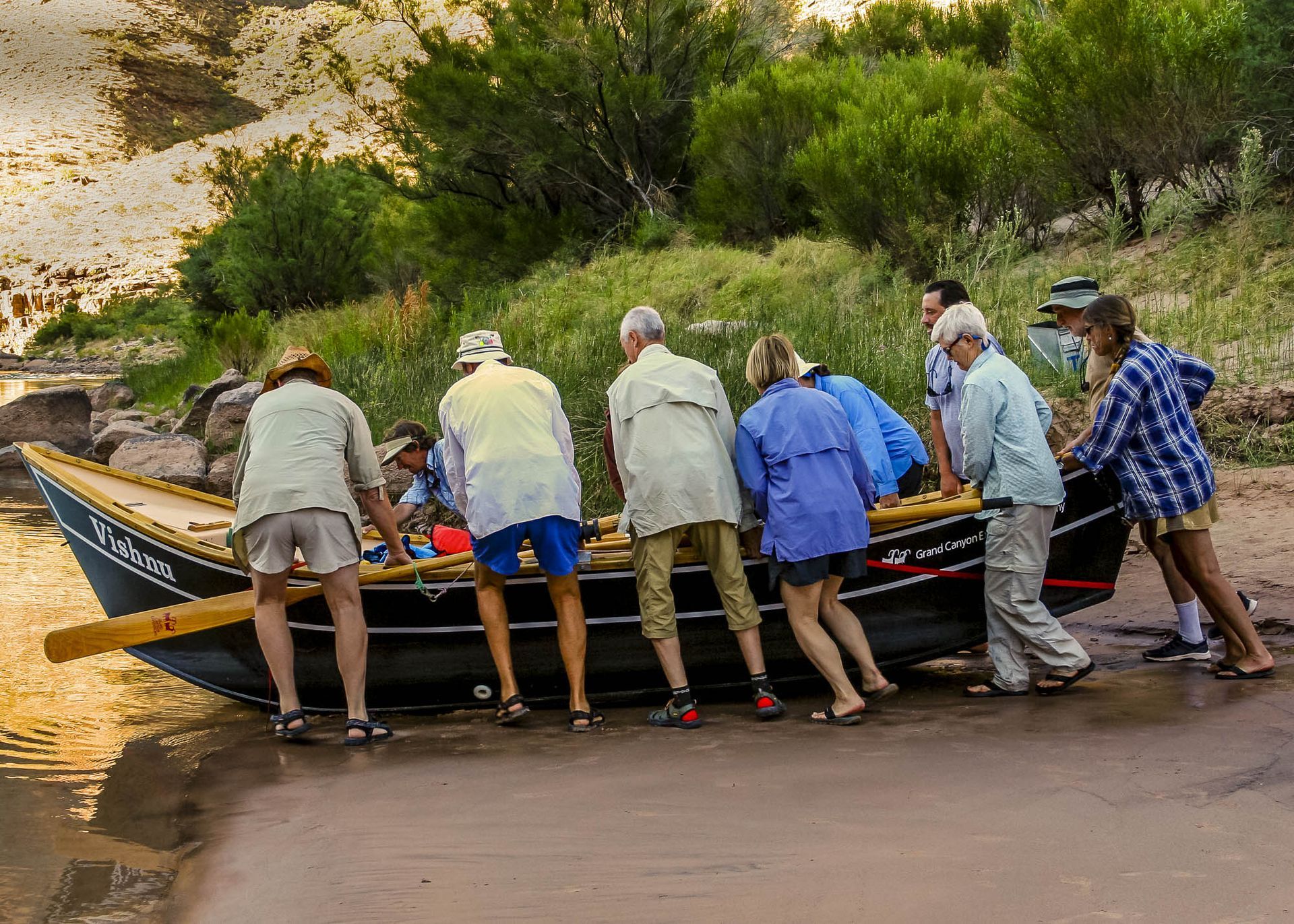 People pushing a wooden boat into a river. Several wear light-colored shirts, shorts. Green foliage and canyon wall in background.