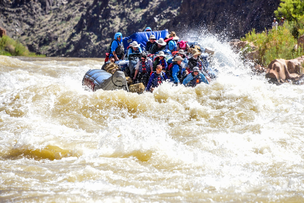 Whitewater raft packed with people surging through rough rapids below rocky canyon walls