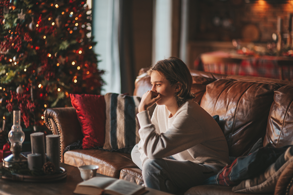 Person sitting on a leather couch in a cozy living room, wrapped in a blanket beside a lit Christmas tree.