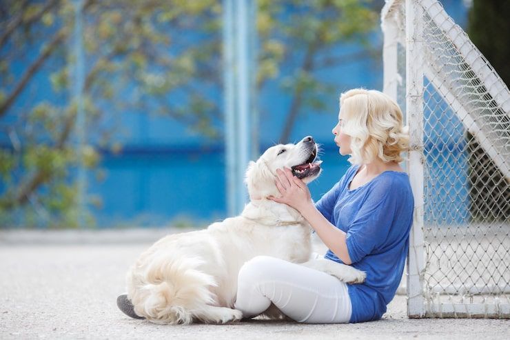 Woman sitting outdoors petting a white dog near a chain-link fence