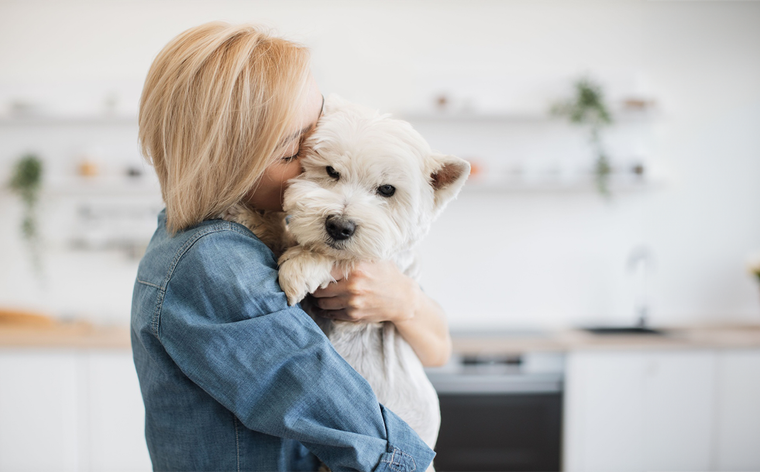 Woman in a denim shirt hugging a small white dog in a bright kitchen