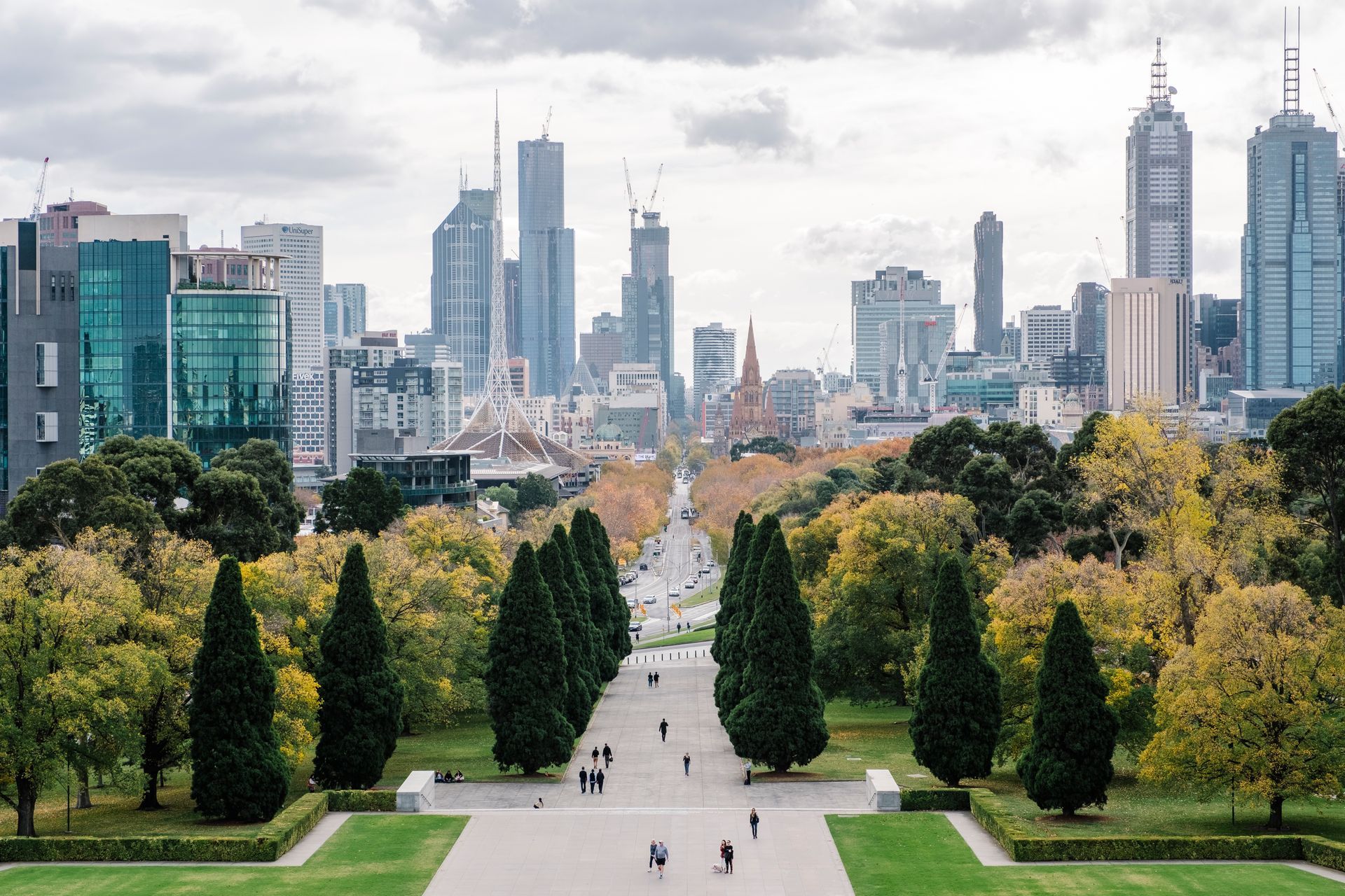 A long, tree-lined path leads toward a skyline of tall buildings under a cloudy sky.