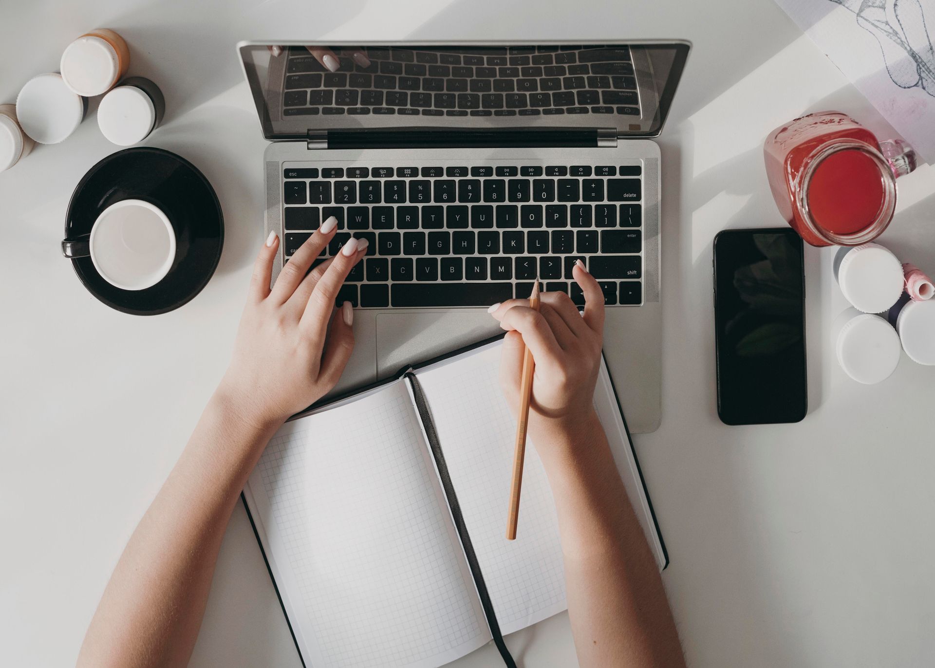 Hands typing on a laptop next to an open notebook and phone on a white desk. A cup of coffee and a drink are also on the desk.