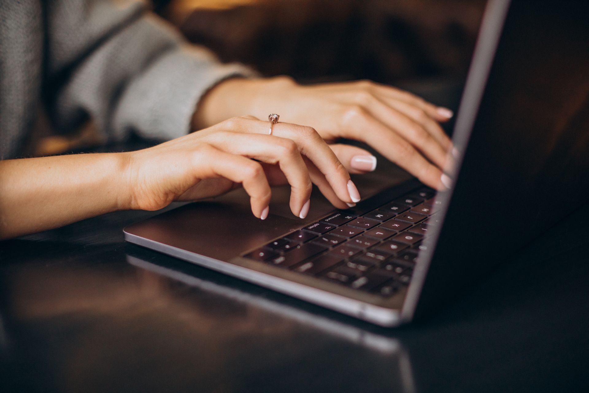 Person typing on a laptop keyboard, hands in focus. A ring is visible on one finger. The background is blurred.