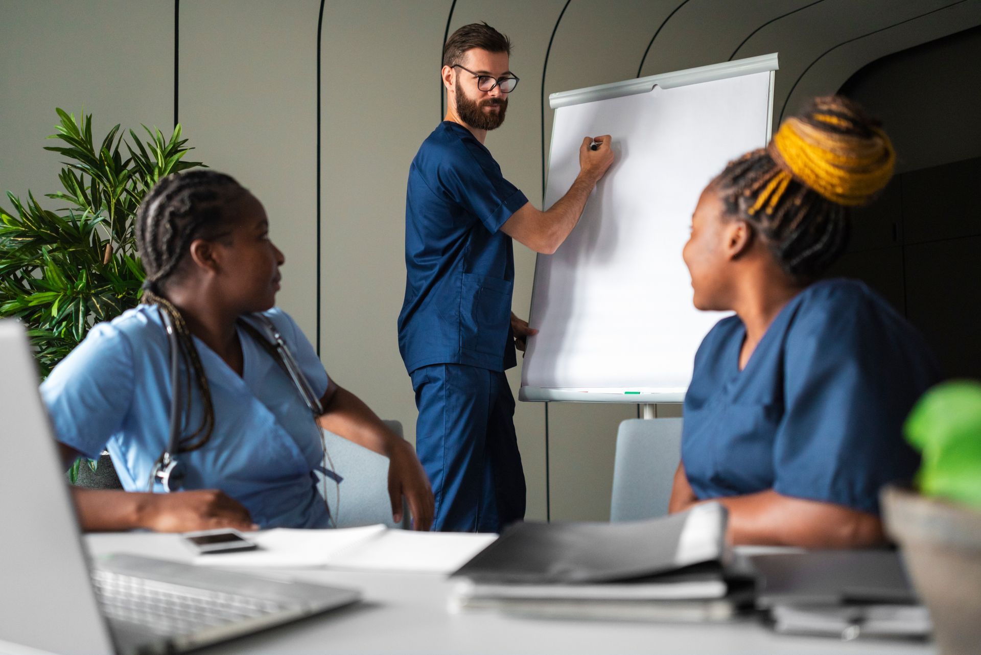 Three healthcare workers in blue scrubs in a meeting; a man writes on a whiteboard as two women listen attentively.