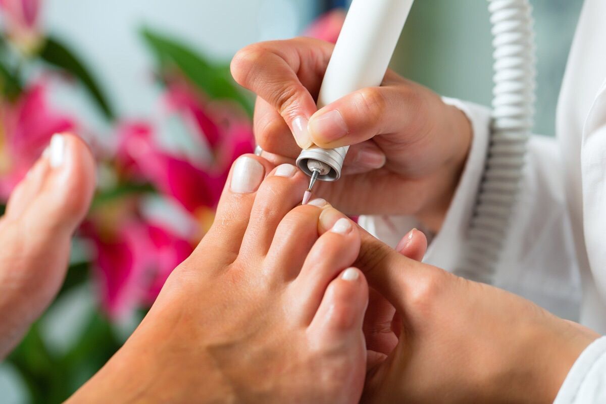 Persona che si fa la pedicure con un trapano bianco, primo piano dei piedi con smalto bianco, fiori rosa sullo sfondo.