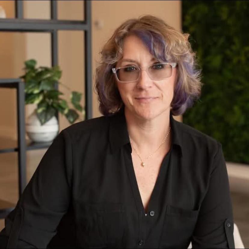 Woman in black shirt and glasses smiles, short hair with purple highlights, in office setting. Woman in black shirt and glasses smiles, short hair with purple highlights, in office setting.