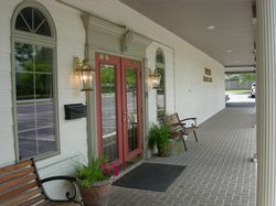A building entrance with red double doors, two arched windows, a covered brick walkway, and wooden benches on each side.