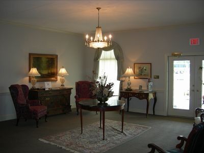 A formal sitting room with a chandelier, two armchairs, side tables with lamps, a central round table, and a glass door.