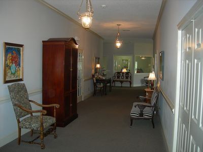 A carpeted indoor hallway features a wooden armoire, a patterned armchair, a small side table, and a bench with seating.
