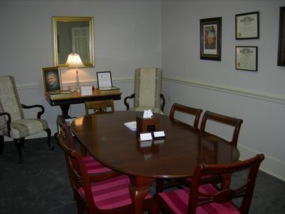 A meeting room with a dark wooden oval table, red striped chairs, a side table with a lamp, and framed art on the walls.
