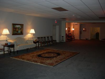 A dimly lit waiting area with a beige sofa, two lamps, a patterned rug, and a row of chairs leading to an inner room.