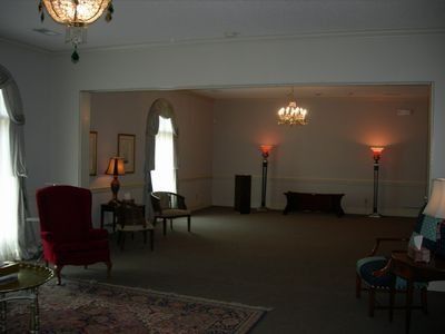 A dimly lit, carpeted room featuring a red armchair, various side tables, and floor lamps under hanging chandeliers.