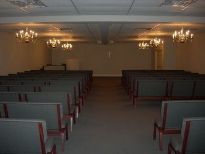 An empty church sanctuary featuring rows of cushioned pews, four chandeliers, and a small cross on the far wall.
