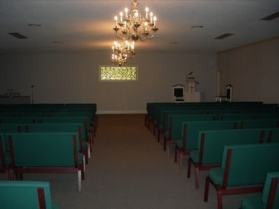 An interior view of a room with rows of green-cushioned wooden pews, two chandeliers, and a small podium at the front.