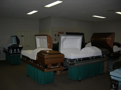 Three open, polished wood caskets displayed on green fabric stands in a dimly lit, white-walled showroom.