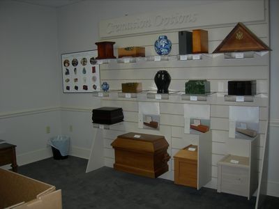 Display of various cremation urns and memorial boxes on tiered shelves in a room with a framed chart on the wall.