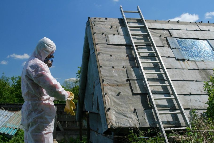 Person in protective suit stands beside a small shed with a ladder leaning against it under a blue sky.