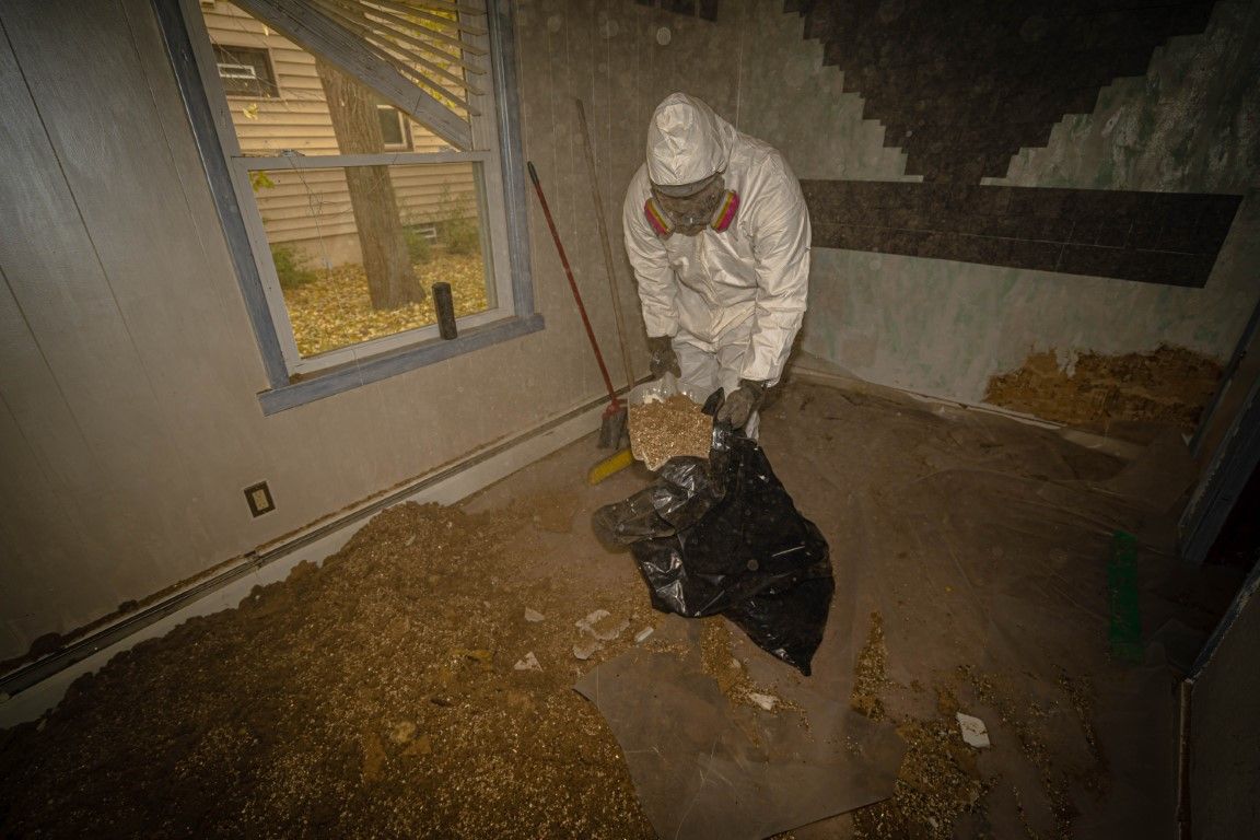 Person in a white suit cleaning debris in a damaged room with a window and scattered insulation.