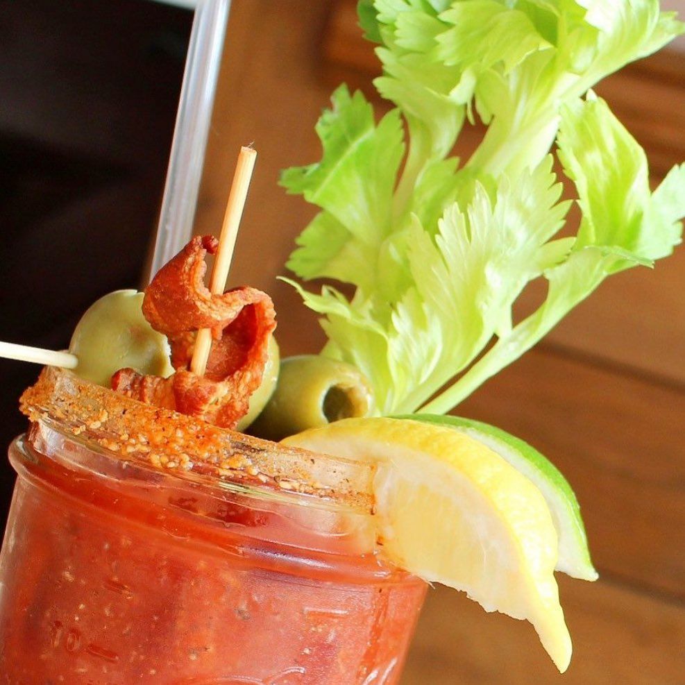 A Bloody Mary cocktail garnished with celery, green olives, a strip of bacon, and a lemon wedge.
